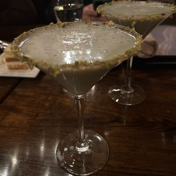 Nu Cavu - Pistachio Martini - Two pistachio martinis with salted rims are positioned on a dark wooden table. The drinks have a creamy, pale green appearance, and the rims are coated with crushed pistachios. In the background, there are glasses of water and people seated, indicating a social dining environment at Nu Cavu restaurant, which offers a rustic dining atmosphere with outdoor views and live entertainment. - Italian Near Me - Wallkill, New York