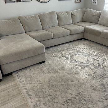 I Clean Carpets - Before - A beige L-shaped sectional sofa with plush cushions is positioned in a living room. The wall behind features decorative clocks and framed artwork, creating a styled interior. The floor is covered with a patterned, neutral-toned area rug, and a window with blinds is partially visible on the right side. - Carpet Cleaning Near Me