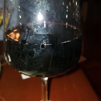 River Mill - Pilot Noir - A close-up of a wine glass filled with red wine, with condensation droplets visible on the exterior of the glass. The background is dark and out of focus, suggesting a low-light environment, possibly a restaurant or bar. The glass is positioned on a wooden table, with a napkin partially visible at the bottom left corner. - New American Near Me - Lynbrook, New York