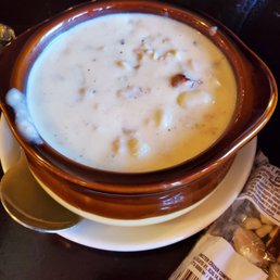New England Clam Chowder - A bowl of clam chowder with a creamy texture and visible chunks of clams, served in a brown ceramic bowl on a white plate.