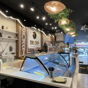 Bilqis Coffee - The image shows the interior of a coffee shop with a dark ceiling and modern lighting fixtures. Hanging woven pendant lights and green plants are suspended from the ceiling, adding decorative elements. The counter area includes a glass display case with ice cream, and the background features shelves with various items and a sign that reads 'BLOIS.' The front entrance is visible with a view of the street outside. - Coffee & Tea Near Me - Brooklyn, New York