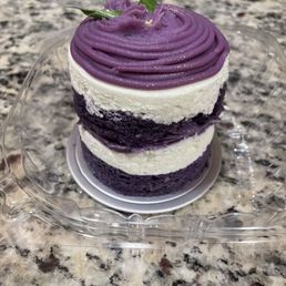 Ube cake - A small, multi-layered cake features alternating purple and white frosting layers. The top is decorated with piped purple frosting in a swirl pattern, garnished with small green leaves. The cake is set on a clear plastic tray on a granite countertop, with a decorative base underneath. The frosting appears smooth and creamy, with a slightly glossy finish.