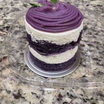 Maprang Bakery & Cafe - Ube cake - A small, multi-layered cake features alternating purple and white frosting layers. The top is decorated with piped purple frosting in a swirl pattern, garnished with small green leaves. The cake is set on a clear plastic tray on a granite countertop, with a decorative base underneath. The frosting appears smooth and creamy, with a slightly glossy finish. - Desserts Near Me - Somerville, Massachusetts
