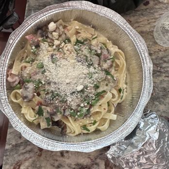 750 Cucina Rustica - Tagliatelle al Bosco - A disposable aluminum container holds pasta topped with grated cheese, chopped herbs, and a creamy sauce, placed on a granite surface. - Italian Near Me - Cary, Illinois