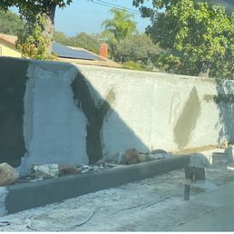 Wall during repair - A partially painted concrete wall with fresh patches and construction supplies in front, set outdoors with trees and rooftops in the background.