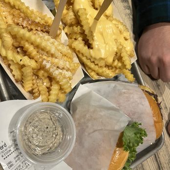 Shake Shack Southdale - A tray holds crinkle-cut French fries with cheese sauce, a small cup of dipping sauce, and a burger wrapped in paper with visible lettuce. The fries are golden and crispy, with some sprinkled with salt. The cheese sauce is creamy and yellow, covering some fries. The dipping sauce appears to be a seasoned or herb-infused sauce. The burger is partially wrapped, showing lettuce and a bun. The tray is on a wooden table, and a person's arm is visible in the background. - Fast Food Near Me - Minneapolis, Minnesota