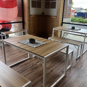 Sharetea - The image shows a cafe interior with natural light coming through large windows. There are wooden tables with white metal frames, each decorated with small potted plants on woven placemats. Benches with matching wooden tops and white frames are positioned along the tables. The floor is wood-patterned, and a corner wall has a wooden panel with bulletin boards. Outside, a red lantern decoration is visible through the window, along with some greenery and a parking lot. - Bubble Tea Near Me - St. Charles, Illinois