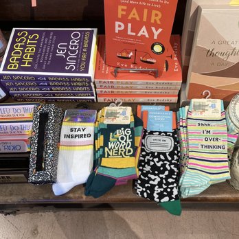 Paper Source - The image shows a store shelf at Paper Source in Pasadena, CA, with a variety of items including books, socks, and a display stand. The books include 'Badass Habits' by Jen Sincero and 'Fair Play' by Eve Rodsky. Several pairs of socks with different patterns and messages such as 'STAY INSPIRED' and 'I'M OVER-THINKING' are arranged on the shelf. The display stand holds multiple copies of 'Fair Play,' a book about relationship management. The items are organized neatly, with some packaging and price tags visible. - Cards & Stationery Near Me - Pasadena, California