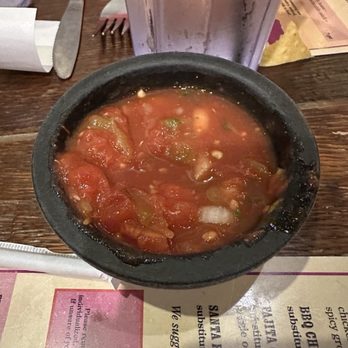 Chile Verde - A black bowl of salsa with visible chunks of tomatoes, onions, and green peppers sits on a wooden restaurant table. Surrounding the bowl are a fork, a knife, a napkin, and a menu. A metallic cup or container is positioned behind the bowl, partially obscuring the view. The table surface is dark wood, and the setting appears to be a casual dining environment. - New Mexican Cuisine Near Me - Columbus, Ohio