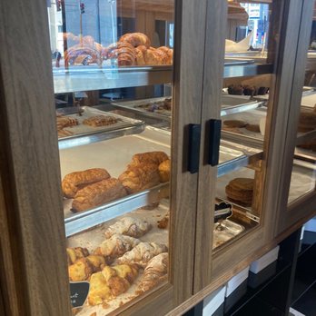 Black Lion Cafe - Pastries - A bakery display case with glass doors contains multiple shelves of baked goods, including croissants, pastries, and other breakfast items. The baked goods are arranged on metal trays, with some items dusted with powdered sugar. The case is made of wood and glass, with black handles on the doors. Through the glass, reflections of the street outside and nearby buildings are visible. - Cafes Near Me - Silver Spring, Maryland
