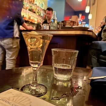 Tonic - Made from the a thought - A wine glass filled with white wine and a glass of water are placed on a wooden table in a bar with patrons and bartenders in the background. - Cocktail Bars Near Me - Santa Fe, New Mexico