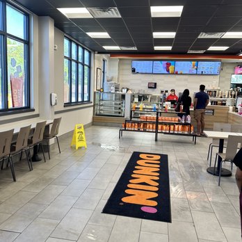 Dunkin' - Inside - The image shows the interior of a Dunkin' Donuts store with large windows on the left side, allowing natural light inside. There are chairs and tables along the windows and a black and orange Dunkin' mat on the tiled floor. The counter area at the back features digital menu screens, staff members, and customers placing orders. A yellow caution sign is on the floor, indicating a wet surface. The ceiling has black tiles with recessed lighting and air vents. - Donuts Near Me - Pocono Summit, Pennsylvania
