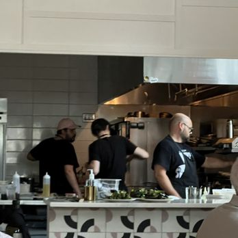 Susan's - Three staff members are working in a restaurant kitchen, with two wearing black shirts and one wearing a pink cap. They are positioned behind a counter with various ingredients and utensils, and are focused on food preparation. The kitchen features stainless steel appliances and a tiled wall, indicating a professional culinary environment. - Salad Near Me - Sawyer, Michigan