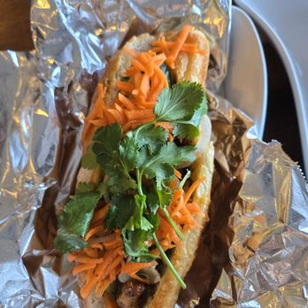 Bamboo by Kulantro - Chicken Banh Mi - A Vietnamese-style banh mi sandwich features grilled meat, shredded carrots, and fresh cilantro inside a baguette. The sandwich is partially wrapped in aluminum foil and is placed on a white plate, with a wooden surface visible in the background. The ingredients are arranged to showcase the toppings and bread, emphasizing the typical components of a banh mi. - Vietnamese Near Me - Albuquerque, New Mexico