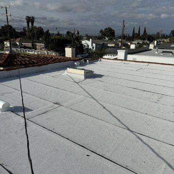 Capitol Roofing - The image shows a flat white roof with vents and a shadow cast by a wire. In the background, there is a residential neighborhood with houses, trees, utility poles, and a cloudy sky overhead. The scene appears to be taken from an elevated perspective, possibly from another roof or high vantage point. - Roofing Near Me - Commerce, California