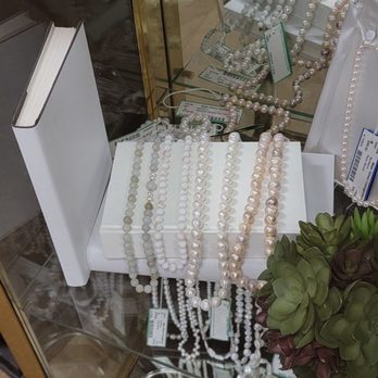 Main Street Consignment - 08152023 - The image shows a collection of pearl necklaces and jewelry arranged on white display stands inside a glass display case. The jewelry includes various styles and colors of pearls, with some necklaces draped over the stands and others lying flat. There are tags and labels attached to some pieces, indicating pricing or product information. A small potted succulent plant is visible in the lower right corner, and the reflection of the jewelry can be seen on the glass surface. - Used, Vintage & Consignment Near Me - Rockwall, Texas