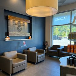 Hand & Stone Massage and Facial Spa - Modern waiting lounge with comfortable gray leather armchairs, large drum pendant light, natural light from windows, and Hand...