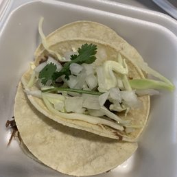 Tacos - A taco filled with shredded meat, topped with chopped onions, cilantro, and shredded cheese, served in a foam takeout container.