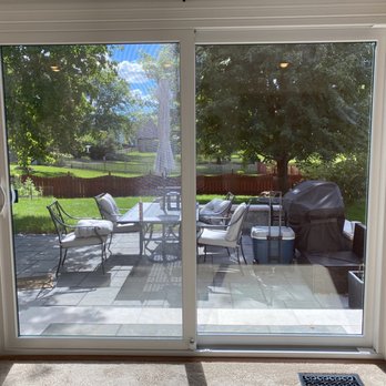 Thermalcraft - Looking our new sliding patio door! - A large sliding glass patio door with white framing provides access to an outdoor backyard patio. The patio features a dining table with chairs, a grill covered with a black cover, and a cooler. The backyard has green grass, trees, and a wooden fence, with a sunny sky and some clouds visible through the trees. The interior floor near the door is carpeted, and a vent is visible at the bottom of the door frame. - Windows Installation Near Me - Villa Park, Illinois