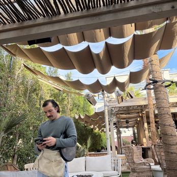Palma - Cabana seating - A man with dark hair and a mustache is standing in an outdoor seating area, looking at his phone. The space features wicker chairs, white cushioned couches, and decorative shade canopies made of fabric and bamboo. There are trees and plants surrounding the area, with a building visible in the background. The setting appears to be a restaurant or cafe with a relaxed atmosphere. - Sushi Bars Near Me - Phoenix, Arizona