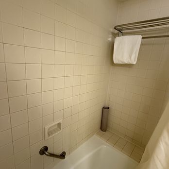 The Clyde Hotel - Standard shower/tub - A shower area features beige ceramic tiles on the walls and a white bathtub. A metal towel rack is mounted on the wall, holding a folded white towel. A shower curtain is partially visible on the right side. There is a small built-in soap dish on the wall and a cylindrical object, possibly a shower rod or pipe, in the corner. - Hotels Near Me - Albuquerque, New Mexico