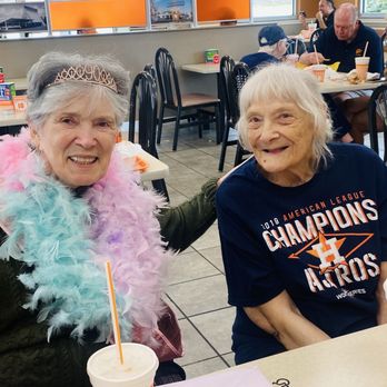 Whataburger - Two elderly women are seated at a table inside Whataburger, smiling at the camera, with drinks in front of them. The woman on the left wears a tiara and a feather boa, while the woman on the right wears a Houston Astros t-shirt. - Burgers Near Me - Tomball, Texas