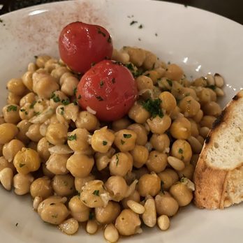 Debajo Tapas ABQ - Garbanzos - A white plate holds a chickpea salad with cooked chickpeas mixed with herbs, garnished with two cherry tomatoes and chopped herbs. A slice of toasted bread is placed on the side of the plate. The dish appears to be served in a restaurant setting. - Spanish Near Me - Albuquerque, New Mexico