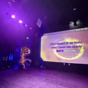 Stage Karaoke x Studios - The image shows an indoor karaoke room with a large screen displaying song lyrics in purple and white text. The room is illuminated by purple and blue stage lighting, creating a vibrant atmosphere. A metallic bunny-shaped balloon is positioned near the wall, adding a decorative element. The floor is tiled, and there is an exit sign visible in the background. The overall setting suggests a lively entertainment space designed for singing and socializing. - Karaoke Near Me - Allston, Massachusetts