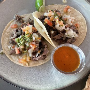 Luna Oaxaca - Carne Asada Tacos - Three carne asada tacos filled with grilled beef, topped with chopped onions, cilantro, and diced tomatoes, served on soft corn tortillas. Lime wedges are placed on the plate, and a small bowl of hot sauce accompanies the dish. The tacos are presented on a gray, round plate, with a casual dining setting. - New Mexican Cuisine Near Me - Baltimore, Maryland