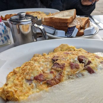 George's - Pastrami omelette - A breakfast plate with an omelette containing bacon pieces, served on a white dish. In the background, there is a plate of toasted bread, a small metal syrup container, and a person wearing a dark jacket sitting at an outdoor table. The setting appears to be a casual diner with outdoor seating. - Diners Near Me - Brooklyn, New York