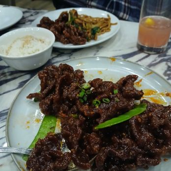 Panda Inn - BEEF...BEEF...BEEF ...D EL I C I O U S N E S S FRFR - A plate of beef in dark sauce with green onions and a green vegetable on a marble table, with rice, noodles, and a drink in the background. - Chinese Near Me - Pasadena, California