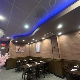 The image shows the interior of a Japanese restaurant with brick walls decorated with artwork. There are wooden tables and chairs arranged for dining, with napkins and utensils set on each table. The ceiling has recessed lighting and blue accent lighting, creating a subdued atmosphere. The space appears clean and organized, with a corner visible that includes a pink decorative element on the left side.