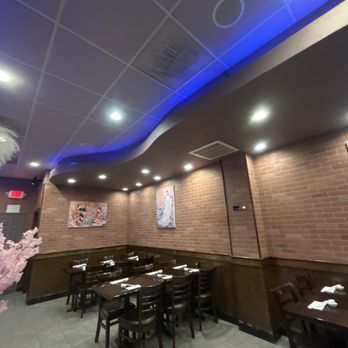 Sakura Grill - The image shows the interior of a Japanese restaurant with brick walls decorated with artwork. There are wooden tables and chairs arranged for dining, with napkins and utensils set on each table. The ceiling has recessed lighting and blue accent lighting, creating a subdued atmosphere. The space appears clean and organized, with a corner visible that includes a pink decorative element on the left side. - Sushi Bars Near Me - Burke, Virginia