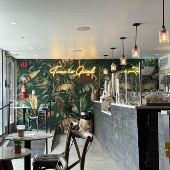 Concept Coffee - Inside - The image shows the interior of a coffee shop with a tropical-themed wall mural of large green leaves and plants. A neon sign reading 'Tune to Grind' is mounted on the wall. Several pendant lights with exposed bulbs hang from the ceiling above the counter. The counter area has coffee-making equipment and supplies, with a glass display case. There are tables and chairs in the seating area, with a small potted plant on one table. An exit sign is visible near the door. - Coffee & Tea Near Me - Queens, New York