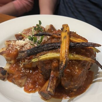 MIX Kitchen & Bar - Pork Osso Buco - A white plate holds pork osso buco topped with roasted carrots and a tomato-based sauce. The dish includes garnishes and appears to be served with a side of bread or similar accompaniment. The setting suggests a restaurant environment, with two people partially visible in the background. - Cocktail Bars Near Me - Ithaca, New York