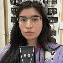 A woman with glasses and long dark hair wears a decorative tiara and holds a jewelry box containing a pearl necklace and matching earrings. She is standing in front of a display wall filled with various jewelry pieces, including earrings and necklaces, in a retail store setting.