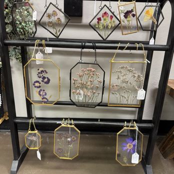 Junk Head - Beautiful, delicate pressed flowers decor - A black metal display stand with three horizontal bars holds pressed flower jewelry in glass frames. The jewelry features different floral arrangements, including daisies, violets, and other wildflowers, with some frames in black or gold-colored metal. The stand is situated indoors, with a wall and some decorative greenery visible in the background. Each piece has a small white or black price tag attached. - Used, Vintage & Consignment Near Me - Mesa, Arizona