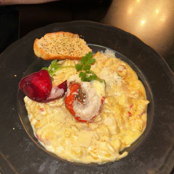 Draft Gastropub - Lobster Mac n cheese, I couldn't recommend this enough. The lobster was divine. The cheese sauce was impeccable.10/10. - A black plate contains lobster mac and cheese topped with shredded cheese and garnished with a sprig of parsley. A lobster claw is placed on top, and a cherry tomato is beside it. A slice of garlic bread is positioned on the edge of the plate. The dish appears creamy with visible pasta and lobster pieces, presented in a casual dining setting. - Gastropubs Near Me - Appleton, Wisconsin