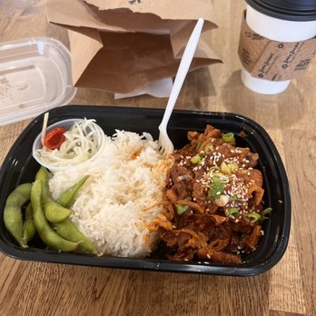 Eggroll Cafe - Korean spicy pork - A takeout meal in a black plastic container includes white rice, green edamame, pickled vegetables with onions and cherry tomato, and spicy pork topped with sesame seeds and chopped green onions. The tray is placed on a wooden table, with a paper bag, plastic container, and coffee cup with a sleeve and lid visible in the background. - Coffee & Tea Near Me - Lowell, Massachusetts