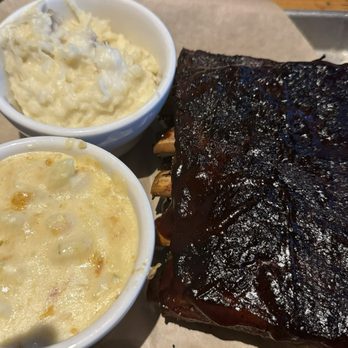 Twin Peaks - Sweet n' Smoky Ribs Adult Mac & Cheese House Made Garlic Mashed Potatoes - A tray holds a serving of smoked ribs with a dark, caramelized exterior, accompanied by two small bowls of mashed potatoes and potato salad, and a small container of barbecue sauce. The meal is presented on a metal tray with parchment paper, set on a wooden table. - American Near Me - Albuquerque, New Mexico