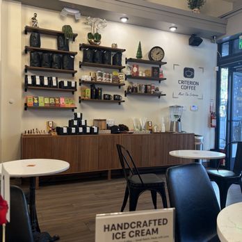 Criterion coffee - The image shows the interior of Criterion Coffee, featuring a wall with multiple shelves holding coffee bags, tea boxes, and decorative objects like plants and a clock. A wooden counter runs along the wall, with various coffee-related items and a beverage dispenser. The seating area includes round tables and black chairs, with natural light coming through the glass door. Potted plants are visible on the upper ledge, contributing to the modern decor. - Coffee & Tea Near Me - Pasadena, California