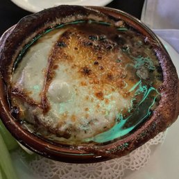French Onion Gratin Soup - A ceramic bowl contains French Onion Soup with melted cheese and toasted bread slices, served on a white doily at a restaurant.
