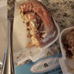Traditional - A large cinnamon roll with icing, partially eaten, sits on a foam plate on a granite countertop. A knife is placed on the plate.