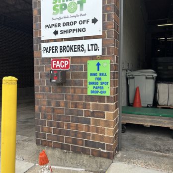 Shred Spot - Sign by back entrance. - Multiple signs are mounted on a brick wall outside Shred Spot in Northbrook, IL. The signs include the business name, directions for paper drop-off and shipping, and instructions to ring a bell for shredding services. A small red FACP box is also visible, along with a yellow bollard and an orange traffic cone on the sidewalk. The background shows part of the business interior with waste bins and a traffic cone. - Recycling Center Near Me - Northbrook, Illinois