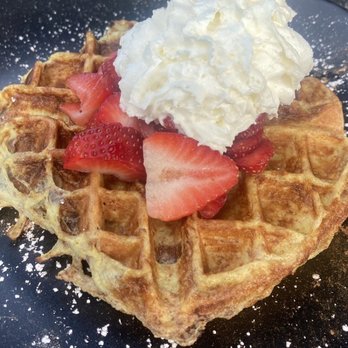 Kitchen 64 - French Toast Waffle w/Strawberry - A heart-shaped waffle topped with sliced strawberries, a dollop of whipped cream, and a dusting of powdered sugar, served on a black plate. - Breakfast & Brunch Near Me - Middlebury, Connecticut
