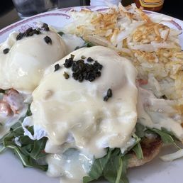 Smoked Salmon Benedicted - A plate features two poached eggs covered in hollandaise sauce, garnished with black pepper, placed on a bed of fresh greens. To the side, there are crispy hash browns with a golden-brown exterior. The dish appears to be a breakfast or brunch meal, served on a decorative plate with a fork visible on the right side.