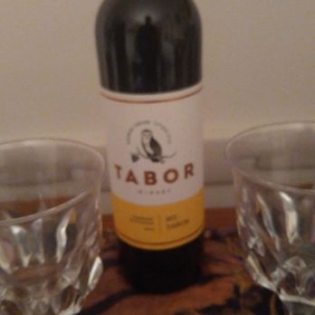 Yankee Spirits - Israeli wine,100% kosher. - A bottle of Tabor wine with a yellow and white label is centered on a wooden surface, flanked by two empty, faceted glasses. The background is plain and neutral, and the image appears slightly blurry. The setting suggests a casual wine-drinking arrangement. - Beer, Wine & Spirits Near Me - Attleboro, Massachusetts