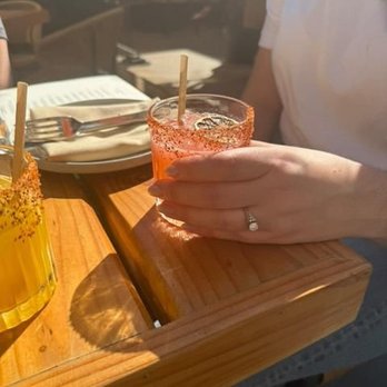 Tabú - A person, wearing a white shirt, is holding a pink cocktail with a straw in a glass, sitting at a wooden table. The table has plates, utensils, and a menu, indicating a dining setting. The background shows a blurred restaurant interior with natural light. - New Mexican Cuisine Near Me - Brooklyn, New York