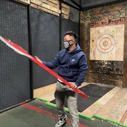 A man in a navy hoodie, gray pants, and sneakers holds a red hockey stick inside an indoor game space with a wooden target and black mesh walls.