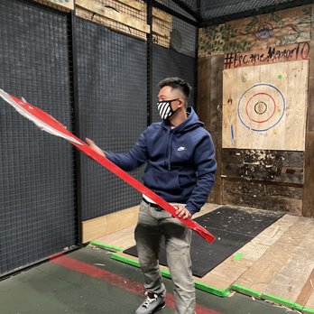 Escape Manor - A man in a navy hoodie, gray pants, and sneakers holds a red hockey stick inside an indoor game space with a wooden target and black mesh walls. - Escape Games Near Me - Toronto, Ontario